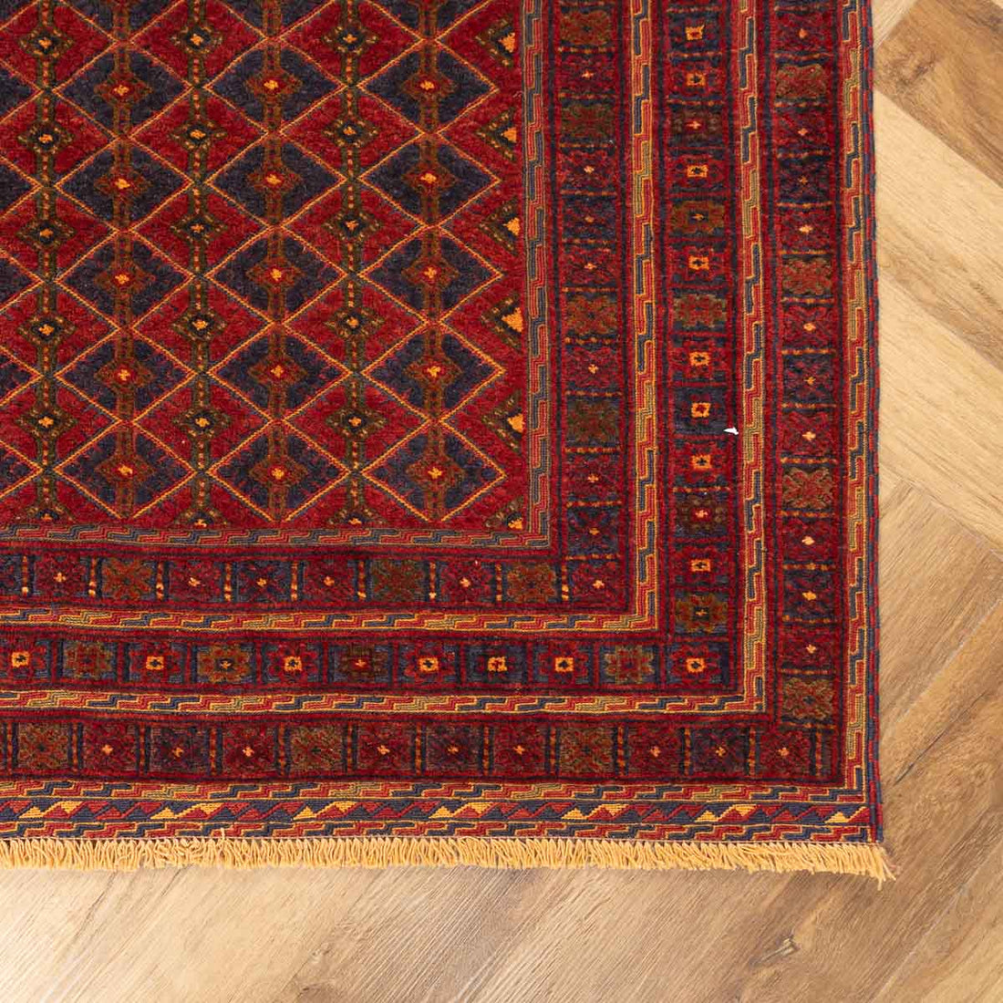 Close-up of the corner of a traditional red and navy Persian-style area rug featuring a repeating diamond pattern, intricate geometric borders, and short fringe, displayed on a light wood floor.