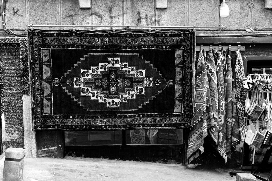 Black and white photo of traditional handwoven rugs displayed in an outdoor market setting.