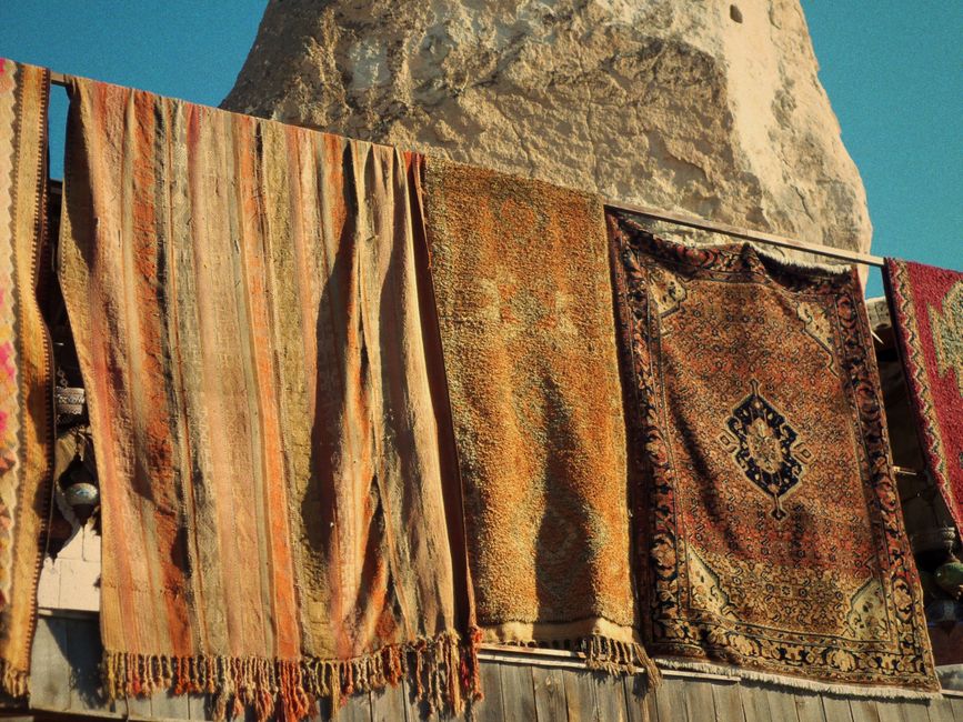 Colorful oriental rugs dry under the sun against a rocky backdrop, showcasing intricate patterns.