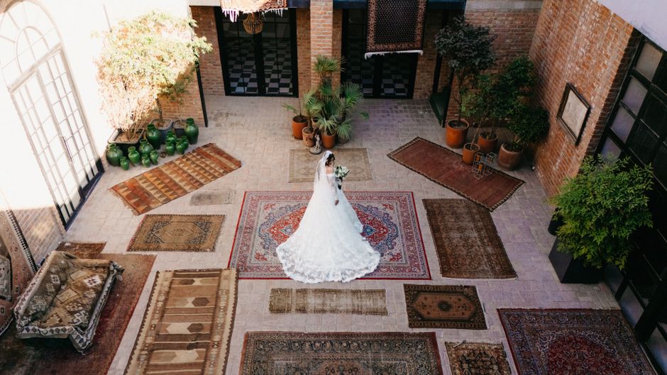 Bride in white gown stands amidst ornate rugs in a sunny courtyard, exuding elegance and charm.